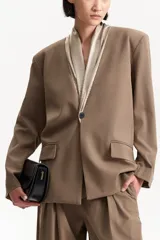 Blazer de color marrón topo, con un solo botón de cierre frontal y solapas anchas con forro interior en tono beige metalizado. Presenta bolsillos de solapa y un corte holgado.