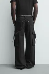 Pantalón cargo de corte relajado en color negro con efecto lavado. Presenta bolsillos laterales tipo cargo con solapas y cintas ajustables, bolsillos frontales y traseros, y detalles de argollas metálicas. Cierre frontal con cremallera y botón.