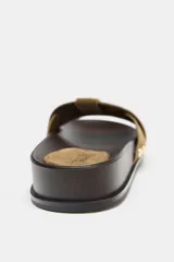 Sandalia plana con tiras cruzadas de gamuza color verde oliva, con detalles de tachas metálicas doradas en los laterales y suela de madera oscura.