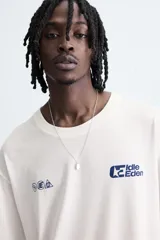 Camiseta de manga corta y cuello redondo, color blanco roto, con efecto de doble capa (la capa inferior gris asoma en el ruedo). Presenta estampados pequeños en azul marino en el pecho: un logo con texto 'Idle Eden' y otros símbolos.