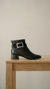 Bota corta de cuero negro con taco cuadrado de 4 cm. Presenta una tira de gamuza en el tobillo con una hebilla metálica rectangular plateada como detalle principal.