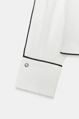 Blusa de crepe con cuello barco y manga larga. Presenta un diseño minimalista en color blanco con detalles de vivos en color negro que recorren el cuello, las mangas y los puños. Los puños incluyen botones decorativos.