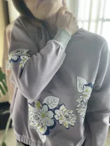 Buzo de algodón color tostado, de corte cropped y estilo oversize. Presenta cuello redondo, mangas largas con puños ajustados y elástico en la cintura. Destaca por una estampa exclusiva de flores en el frente y en una de las mangas.
