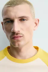 Camiseta de corte relajado con mangas raglán cortas en color amarillo y cuerpo en color crema. Presenta un estampado gráfico frontal con temática de campamento de básquetbol y un cocodrilo.