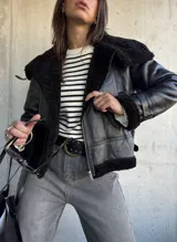Campera tipo gamulán de cuero sintético negro con interior y detalles de cuello y puños en piel sintética negra. Presenta cierre frontal, correas ajustables en los puños y diseño de corte clásico.