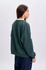 Sudadera polo verde oscuro con cuello y franja central en color beige. Presenta un diseño de manga larga y cuenta con un bordado frontal con el texto "BROOKLYN ATHLETICS DEPT. Est. 1995".