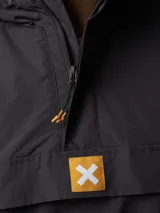 Canguro tipo anorak de color gris oscuro, con capucha ajustable, bolsillo frontal con solapa y cierre de media cremallera. Presenta detalles en color naranja en los tiradores de los cierres y un parche con el logo de la marca en el centro del pecho.