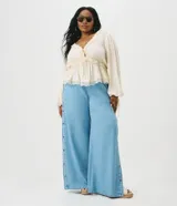 Blusa de viscosa texturada color marfil, con escote en V pronunciado, lazo frontal y corte estilo túnica. Presenta mangas largas abullonadas con puños ajustables mediante tiras y detalles de encaje en el escote, la cintura fruncida y el ruedo.