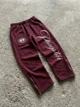 Pantalón de jogging bordó con diseño estampado en blanco. Presenta un logo circular con el número 27 en la pierna derecha y la palabra Plagstar en tipografía cursiva en la pierna izquierda. Cuenta con cierres metálicos en los tobillos.