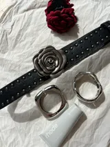 Cinturón negro de cuero sintético con remaches plateados y hebilla en forma de rosa plateada.