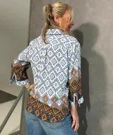 Blusa de manga larga con cuello camisero, corte recto y cierre frontal de botones. Presenta un diseño estampado geométrico en tonos celeste, blanco y marrón.