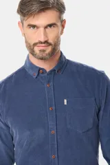 Camisa de pana fina en color verde oscuro, de manga larga con puños arremangados, cuello camisero con botones y un bolsillo aplicado en el pecho con pequeña etiqueta decorativa.