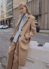 Abrigo largo de corte recto confeccionado en tejido mezcla de lana color camel. Presenta cuello con solapas de muesca, cierre frontal de doble botonadura, manga larga, un bolsillo de ribete en el pecho y dos bolsillos con solapa en la parte frontal. Incluye forro interior y abertura posterior en el bajo.