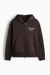 Campera canguro de felpa marrón oscuro, con cierre frontal completo, capucha, bolsillos delanteros tipo parche y pequeño bordado blanco en el pecho con texto y estrellas.
