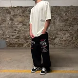 Pantalón de jean negro de corte super baggy con bordados de logos de marcas de automovilismo y texto en blanco y rojo.