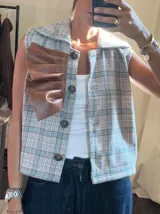 Chaleco de corte recto con diseño a cuadros en tonos beige, azul y marrón. Presenta cuello camisero, cierre frontal con botones y un detalle decorativo de volado en cuero sintético color marrón sobre el lado derecho del pecho.