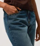 Pantalón de jean corte bootcut con tiro bajo, confeccionado en denim de algodón con lavado dirty que aporta un efecto envejecido. Presenta cierre frontal con botón y cremallera, presillas en la cintura y detalle de tachas metálicas en los bolsillos frontales.