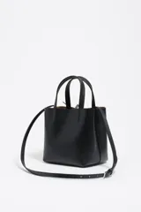 Bolso pequeño de piel vacuna color negro, con estructura rígida, doble asa de mano y correa larga ajustable para llevar al hombro o cruzado. Presenta cierre de botón a presión y logo metálico de la marca en el frente.