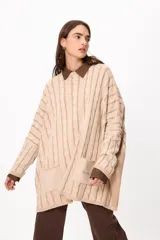 Sweater de punto con diseño de trenzas verticales, color crudo y camel, con bolsillos frontales y detalle de logo bordado. Presenta un corte holgado y mangas largas.