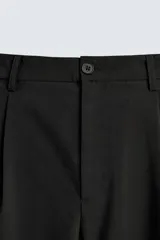 Pantalón negro de corte ancho con pliegues frontales dobles, bolsillos delanteros y traseros de vivo. Cierre frontal con cremallera y botón.