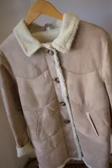 Campera tipo gamulán de cuero genuino en color marrón, con interior y detalles en corderito sintético color crema. Presenta cierre frontal con botones, cuello solapa y puños con vuelta.