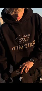Hoodie de color negro con diseño de estampa frontal que combina una tipografía manuscrita con el texto ITTAI STARS en letras mayúsculas. Presenta un corte holgado, capucha amplia y bolsillo tipo canguro.