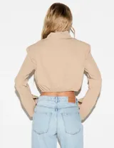 Camisa crop beige de manga larga con cuello alto abotonado, hombreras y puños amplios. Tiene terminación elastizada en la cintura y cierre frontal de botones.