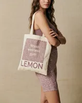 Bolsa tipo tote bag color crudo, con tiras de hombro, confeccionada en 100% algodón. Presenta un diseño gráfico frontal con estampado en tonos morados que incluye texto con las palabras "fitness, wellness, gym" y la marca "LEMON" en la parte inferior.