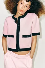 Chaqueta de punto rosa con ribetes negros en el cuello, la botonera, los bolsillos, los puños y el ruedo. Cuenta con manga corta, cuello redondo, bolsillos frontales de plastrón con botones y cierre frontal abotonado.
