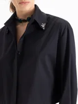 Camisa de popelina de algodón con corte holgado y largo a la cintura. Presenta cierre frontal con botones ocultos, mangas largas con puños abotonados y detalle de punteras metálicas en las puntas del cuello.