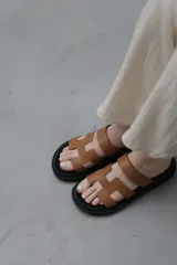 Sandalias planas de cuero sintético, con base negra y tiras anchas en forma de H en color caramelo o negro.