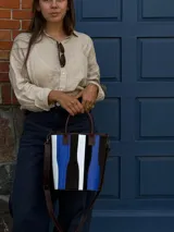 Matera de cuero con diseño de paneles verticales en combinación de colores azul, verde, marrón y bordó. Cuenta con dos asas cortas de cuero y estructura rígida.