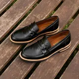 Mocasín de cuero vacuno negro con textura grabada tipo reptil. Presenta un diseño clásico con antifaz en el empeine, suela de goma en tono natural y taco bajo de madera.