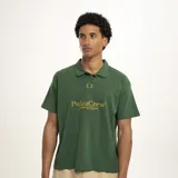 Remera tipo polo de manga corta, color verde oscuro, con cuello y abertura frontal. Presenta un estampado dorado en el pecho con el texto "PulauCrew" y un pequeño logo de corona de laurel bordado.