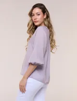 Blusa de gasa de color lila con calce holgado, cuello a la base con escote en V y cartera de botones a tono. Cuenta con fruncidos en el cuello y hombros que otorgan volumen, manga tres cuartos estilo abullonado con puño elastizado y ruedo suavemente redondeado.
