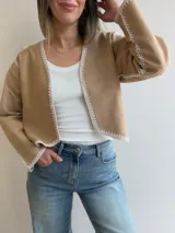 Chaqueta de efecto gamuza color tostado, con diseño abierto sin cierres y detalles de costuras artesanales tipo crochet en color blanco en los bordes, el frente, el cuello y las mangas.