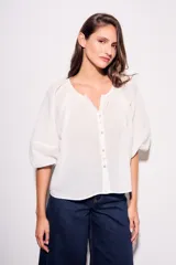 Blusa blanca de algodón peinado, con escote redondo y abertura frontal con botones. Presenta mangas globo de tres cuartos con elástico en el puño y corte holgado.