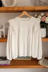 Camisa blanca de manga larga con detalles de volados en los hombros y bordado geométrico en el pecho. Presenta un diseño holgado con puños elásticos.