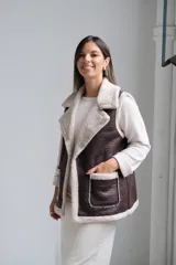 Chaleco de cuero sintético color chocolate con interior y bordes de corderito beige. Presenta solapas amplias, dos bolsillos frontales con detalle de corderito y corte recto.
