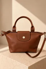 Cartera petit de cuero vacuno color suela, con manija superior y correa larga desmontable. Cuenta con cierre principal y solapa frontal con detalle de botón metálico grabado.