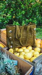 Bolso estilo shopper de cuero con textura efecto cocodrilo en color verde oliva. Presenta detalles de flecos laterales, remaches metálicos circulares y doble asa de hombro.