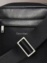 Cartera reporter estructurada Calvin Klein de poliuretano con textura granulada, color negro, cierre superior de cremallera y bolsillo frontal con cierre. Cuenta con correa de hombro ajustable y logo metálico de la marca en la parte delantera.