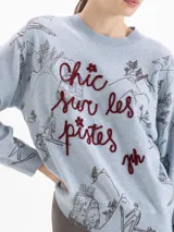 Sweater de lana color celeste con silueta holgada, cuello redondo y mangas largas. Presenta un estampado integral de paisajes de montaña y esquí, con la frase 'Chic sur les pistes' bordada en relieve en color bordó.
