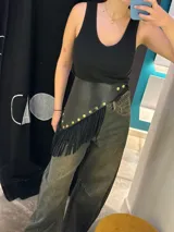 Cinto de cuero negro con diseño asimétrico, decorado con tachas metálicas doradas y flecos largos en el borde inferior.