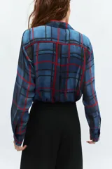 Camisa de manga larga con cuello solapa y cierre frontal con botones. Presenta un diseño de cuadros en tonos azules y rojos sobre un tejido semitransparente.