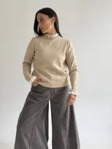 Sweater de punto color beige, con cuello redondo adornado con apliques de pedrería y detalle de puntilla blanca en el cuello y los puños.