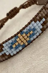 Pulsera tejida con cuentas Miyuki, con diseño geométrico en tonos marrón, celeste, azul y dorado. Cuenta con un sistema de cierre ajustable mediante cordón marrón.