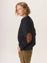 Chaqueta de paño color marrón oscuro, de corte clásico y cuello redondo, con cierre frontal dorado. Presenta bolsillos rectangulares con ribetes en color rojizo y parches de pana a rayas en los hombros.