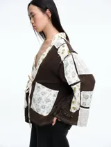 Camisa de estilo kimono con diseño patchwork, confeccionada en una combinación de tela texturizada color marrón y paneles de tela estampada con motivos florales en tonos claros. Presenta cierre frontal mediante lazo, mangas largas y bolsillos frontales.