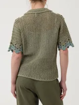 Sweater verde de tejido tipo crochet, con cuello camisero, mangas cortas y cierre frontal de botones. Presenta un diseño de bandas laterales con aplicaciones florales en tonos blancos y celestes.